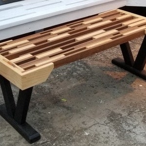 May include: A wooden bench with a mosaic-style top made of various wood tones. The bench has black, V-shaped legs and light-colored wood accents on the ends. The bench is approximately 48 inches long.