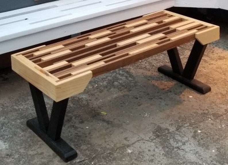 May include: A wooden bench with a mosaic-style top made of various wood tones. The bench has black, V-shaped legs and light-colored wood accents on the ends. The bench is approximately 48 inches long.