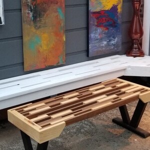 May include: A wooden bench with a mosaic-style top made of various wood tones. The bench has black V-shaped legs and light-colored wood accents on the ends. Two abstract paintings hang in the background.