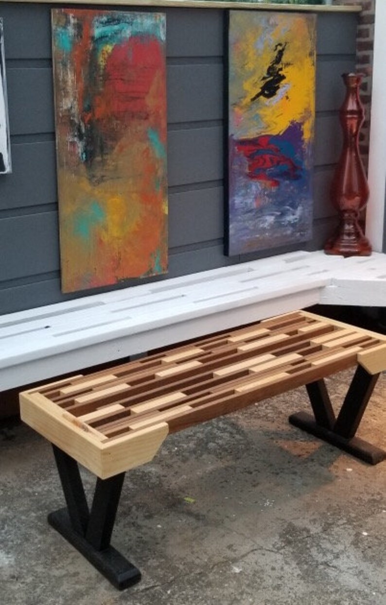 May include: A wooden bench with a mosaic-style top made of various wood tones. The bench has black V-shaped legs and light-colored wood accents on the ends. Two abstract paintings hang in the background.