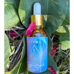 Puede incluir: Una pequeña botella de vidrio de aceite facial Rose-Kissed con una tapa dorada y un cuentagotas blanco. La etiqueta es azul con texto blanco y un diseño de gota de agua. La botella está rodeada de hojas verdes y pétalos de flores rosas.