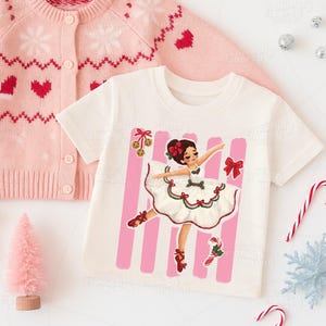 Puede incluir: Una camiseta de manga corta color crema con un gráfico de una bailarina con vestido blanco y ribetes rojos y verdes, rodeada de rayas verticales rosas, un lazo rojo, un bastón de caramelo y campanas doradas, junto a un cárdigan de punto rosa.