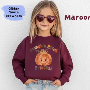 May include: A maroon Gildan youth crewneck sweatshirt featuring a graphic of a plaid pumpkin wearing a crown with the text Pumpkin Patch Princess. The sweatshirt has long sleeves and a ribbed neckline, presented in a solid dark red color.
