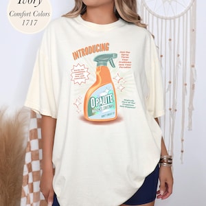 May include: Ivory-colored t-shirt with a graphic of an orange cleaning spray bottle labeled "OPALITE MAGICAL RESULTS." The shirt also features text that reads "INTRODUCING" and "Just One Spray Turns Your Problems Into Your Paradise."