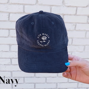 May include: Navy blue corduroy baseball cap with the text "AVE MARIA GRATIA PLENA" in a circular design. The design includes a rose graphic. The cap is a classic six-panel style with a curved brim.