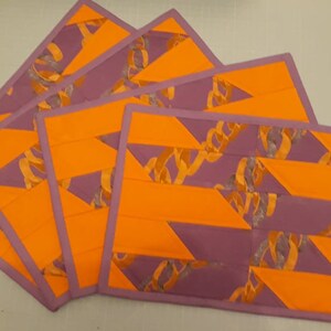 May include: Four orange and purple placemats with a geometric pattern. The placemats have a purple border and a geometric pattern of orange and purple stripes.
