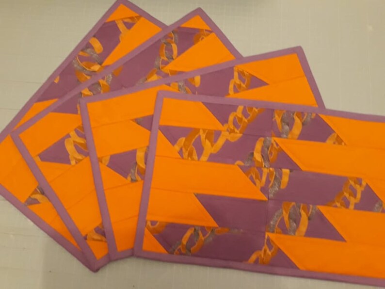 May include: Four orange and purple placemats with a geometric pattern. The placemats have a purple border and a geometric pattern of orange and purple stripes.
