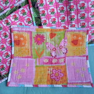 May include: Set of four pink and green quilted placemats with a floral pattern. The placemats have a white border with a blue and pink trim. One placemat features a butterfly and flower design.