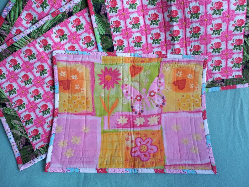May include: Set of four pink and green quilted placemats with a floral pattern. The placemats have a white border with a blue and pink trim. One placemat features a butterfly and flower design.