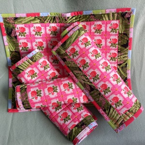 May include: Set of four pink and green quilted placemats with a floral pattern. The placemats are rolled up and feature a border of green asparagus.