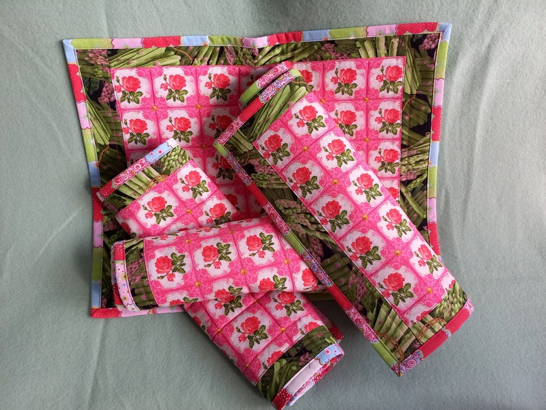 May include: Set of four pink and green quilted placemats with a floral pattern. The placemats are rolled up and feature a border of green asparagus.