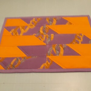 May include: A purple and orange patchwork quilt with a geometric pattern. The quilt is made of different fabrics and has a unique design.