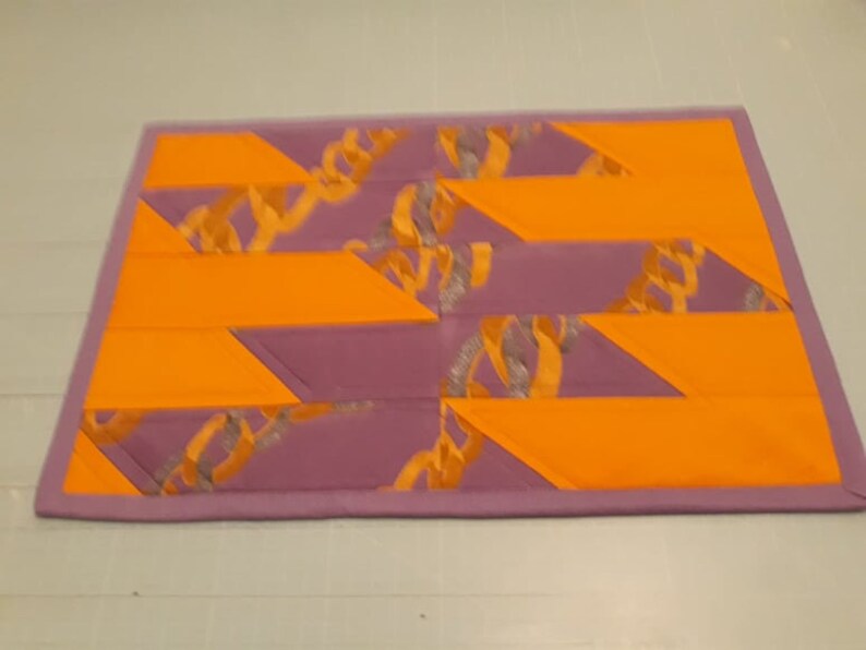 May include: A purple and orange patchwork quilt with a geometric pattern. The quilt is made of different fabrics and has a unique design.