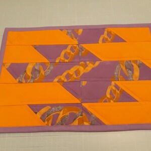 May include: A purple and orange quilted placemat with a geometric pattern of interlocking triangles. The fabric features a gold chain design.