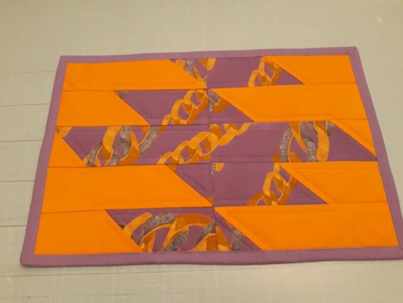 May include: A purple and orange quilted placemat with a geometric pattern of interlocking triangles. The fabric features a gold chain design.
