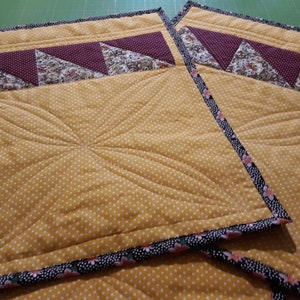 K&ouml;nnte beinhalten: Zwei gelbe gequiltete Platzsets mit einem schwarzen und rosafarbenen Blumensaum. Die Platzsets haben ein gestepptes Design und ein burgunderfarbenes und florales Stoffpanel oben.