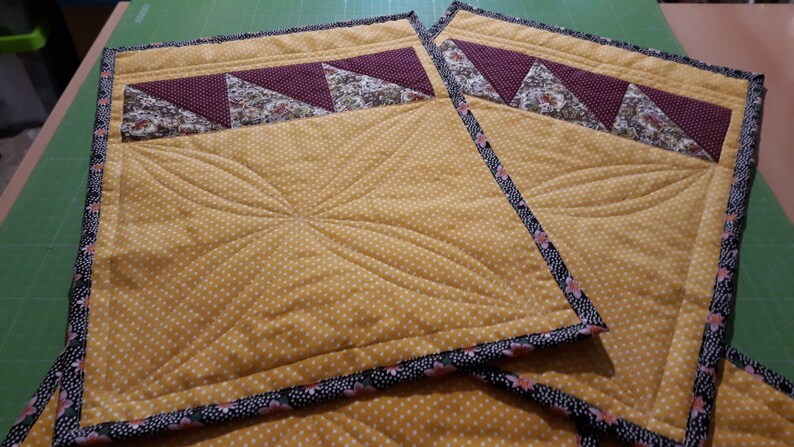 K&ouml;nnte beinhalten: Zwei gelbe gequiltete Platzsets mit einem schwarzen und rosafarbenen Blumensaum. Die Platzsets haben ein gestepptes Design und ein burgunderfarbenes und florales Stoffpanel oben.