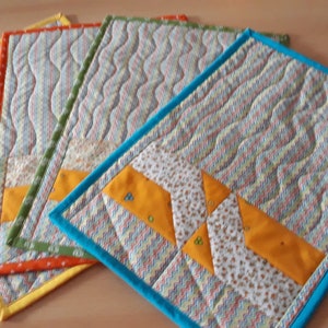 Könnte beinhalten: Vier gequiltete Tischsets mit verschiedenen farbigen Rändern. Die Tischsets sind aus hellblauem Stoff mit einem wellenförmigen Muster. Die Ränder sind gelb, orange, grün und blau.