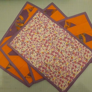 May include: Four purple and orange fabric placemats with a floral pattern. The placemats are layered on top of each other.