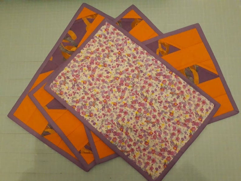 May include: Four purple and orange fabric placemats with a floral pattern. The placemats are layered on top of each other.