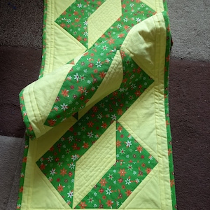 Könnte beinhalten: Ein grüner und gelber Tischläufer mit geometrischem Muster. Der Läufer ist aus Stoff mit Blumenmuster.