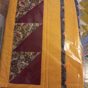 K&ouml;nnte beinhalten: Ein gesteppter Tischl&auml;ufer mit gelbem Hintergrund und einem burgunderfarbenen und floralen Muster. Der L&auml;ufer hat einen schwarzen und wei&szlig;en Besatz mit kleinen floralen Akzenten.