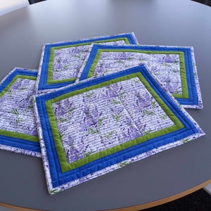 Könnte beinhalten: Vier gequiltete Platzsets mit einem Lavendel-Blumenmuster und blauem und grünem Besatz. Die Platzsets liegen auf einem grauen Tisch.