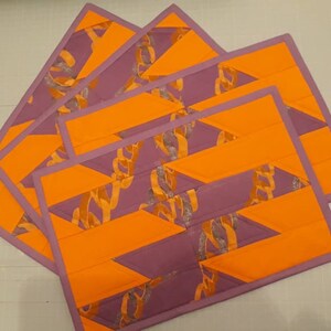 May include: Set of four purple and orange quilted placemats with a geometric pattern. The placemats are made of fabric with a silver glitter accent.