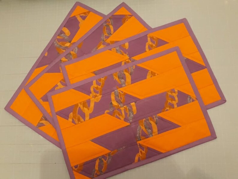 May include: Set of four purple and orange quilted placemats with a geometric pattern. The placemats are made of fabric with a silver glitter accent.