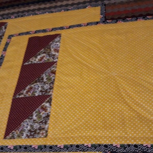 K&ouml;nnte beinhalten: Zwei gelbe Stoff-Platzsets mit einem burgunderfarbenen und braunen geometrischen Muster. Die Platzsets haben einen schwarzen und wei&szlig;en Rand.