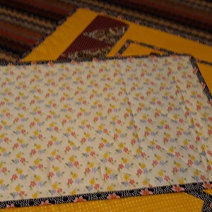 K&ouml;nnte beinhalten: Eine Steppdecke mit einem Blumenmuster auf wei&szlig;em Hintergrund. Die Steppdecke ist mit einem schwarzen und wei&szlig;en Besatz eingefasst. Die Steppdecke liegt auf einem gelben Stoff mit einem kleinen Punkte-Muster.