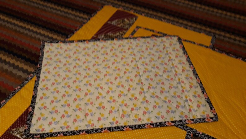 K&ouml;nnte beinhalten: Eine Steppdecke mit einem Blumenmuster auf wei&szlig;em Hintergrund. Die Steppdecke ist mit einem schwarzen und wei&szlig;en Besatz eingefasst. Die Steppdecke liegt auf einem gelben Stoff mit einem kleinen Punkte-Muster.