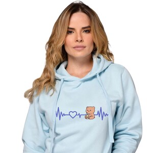Puede incluir: Sudadera con capucha azul claro con cordón y un diseño gráfico de línea de latidos del corazón con un corazón y un oso de dibujos animados bebiendo una bebida. El oso es de color marrón claro.