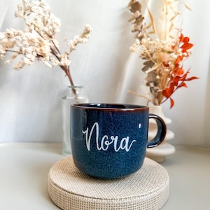 Könnte beinhalten: Ein dunkelblauer Keramikbecher mit gesprenkelter Oberfläche und einem Henkel auf der rechten Seite. Der Name Nora ist in weißer Schreibschrift auf der Vorderseite des Bechers zu sehen, ergänzt durch ein kleines weißes Herzsymbol rechts neben dem Namen.