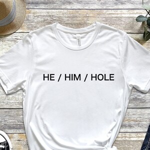 以下が含まれることがあります： 前面に黒い文字で「HE / HIM / HOLE」とプリントされた白いTシャツです。