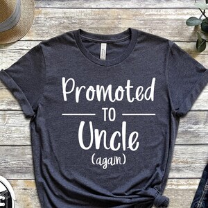 May include: A dark gray t-shirt with the text "Promoted to Uncle (again)" in white lettering.
