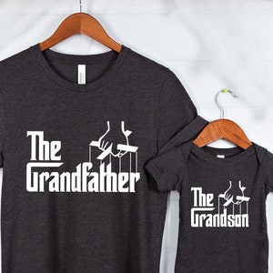 May include: A dark gray t-shirt with the text "The Grandfather" in white with a puppet master hand graphic. A matching dark gray baby bodysuit with the text "The Grandson" in white with a puppet master hand graphic.