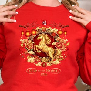 May include: A red sweatshirt featuring a golden horse design within a circular frame, surrounded by flowers and lanterns. The text "Year of the Horse 2026" is displayed below the horse. The sweatshirt is part of a Lunar New Year theme.