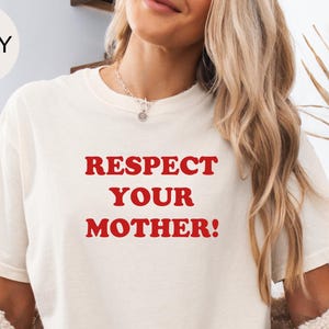 May include: An ivory short-sleeved t-shirt featuring the text "RESPECT YOUR MOTHER!" printed in bold red capital letters across the chest. The shirt has a crew neckline and a relaxed fit, paired with light blue denim shorts and a textured cardigan.