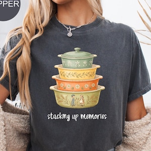 May include: A charcoal gray short-sleeve t-shirt featuring a graphic of four stacked vintage-style mixing bowls in shades of green, yellow, and orange with floral patterns and a small illustration of two children, with the text stacking up memories.