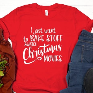May include: A red t-shirt with the text "I just want to BAKE STUFF & WATCH Christmas MOVIES" in white. The shirt is paired with blue jeans, black shoes, and a brown scarf with green foliage.