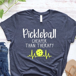 Może przedstawiać: Koszulka w kolorze granatowym z napisem "Pickleball Cheaper Than Therapy" i ż&oacute;łtym obrazkiem piłki do pickleballa z linią bicia serca.