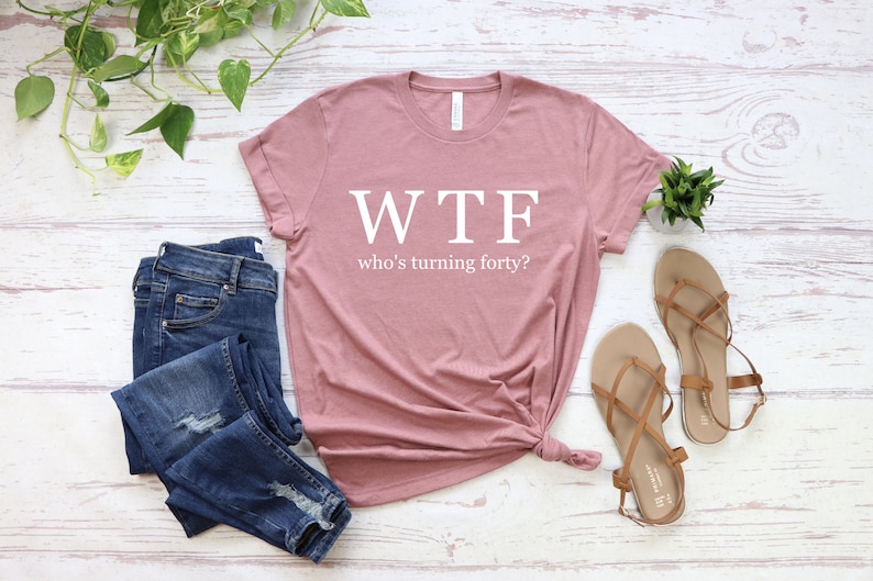 K&ouml;nnte beinhalten: Ein staubiges rosafarbenes T-Shirt mit dem Text "WTF who's turning forty?" in Wei&szlig;. Das Shirt liegt flach auf einer wei&szlig;en Holzoberfl&auml;che mit einem Paar braunen Sandalen und einer blauen Jeans.