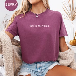Op de afbeelding: Bessenkleurig T-shirt met de witte tekst "dibs on the villain". Het shirt heeft korte mouwen en een ronde hals. De afbeelding toont ook een spijkerbroekshort en een cr&egrave;mekleurig vest.