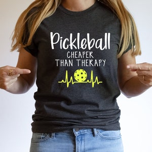 Może przedstawiać: Szary t-shirt z napisem "Pickleball Cheaper Than Therapy" w kolorze białym. Ż&oacute;łta piłka do pickleballa z czarnymi szwami znajduje się w białym wzorze bicia serca.