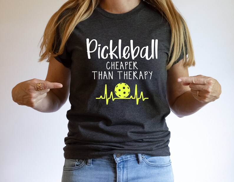 Może przedstawiać: Szary t-shirt z napisem "Pickleball Cheaper Than Therapy" w kolorze białym. Ż&oacute;łta piłka do pickleballa z czarnymi szwami znajduje się w białym wzorze bicia serca.