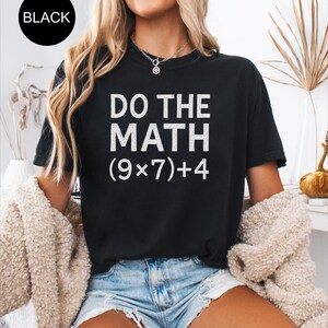 May include: Black t-shirt with the text "DO THE MATH (9x7)+4" in white. The shirt is a crew neck style and is being worn with denim shorts. A black circle with the word "BLACK" is in the upper left corner.