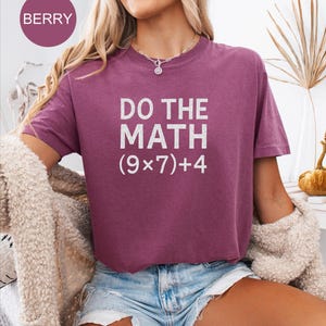 May include: A berry-coloured t-shirt with the text "DO THE MATH (9x7)+4" in white. The shirt is short-sleeved and has a crew neck. The model is wearing denim shorts and a cream-coloured cardigan.