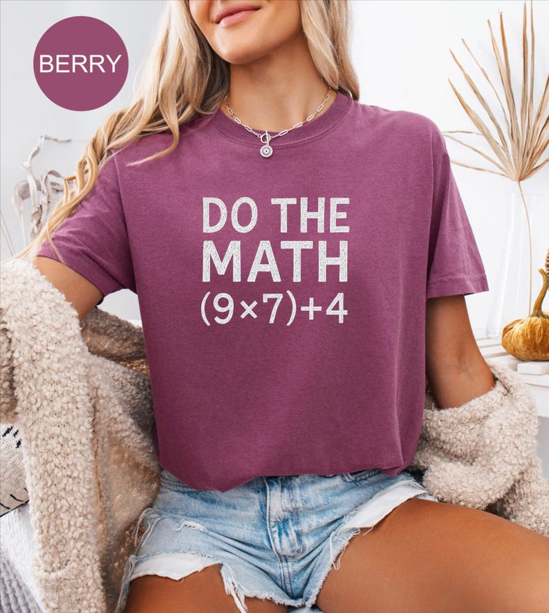 May include: A berry-coloured t-shirt with the text "DO THE MATH (9x7)+4" in white. The shirt is short-sleeved and has a crew neck. The model is wearing denim shorts and a cream-coloured cardigan.