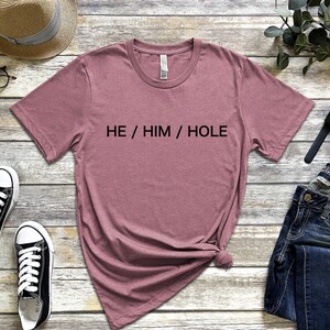以下が含まれることがあります： 前面に「HE / HIM / HOLE」の文字が印刷された、薄紫色のTシャツです。